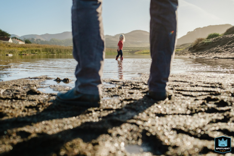 In San Francisco, California, a professional documentary-style photographer captures a daughter standing in a pond, framed between her dad's legs, as she looks back at him.